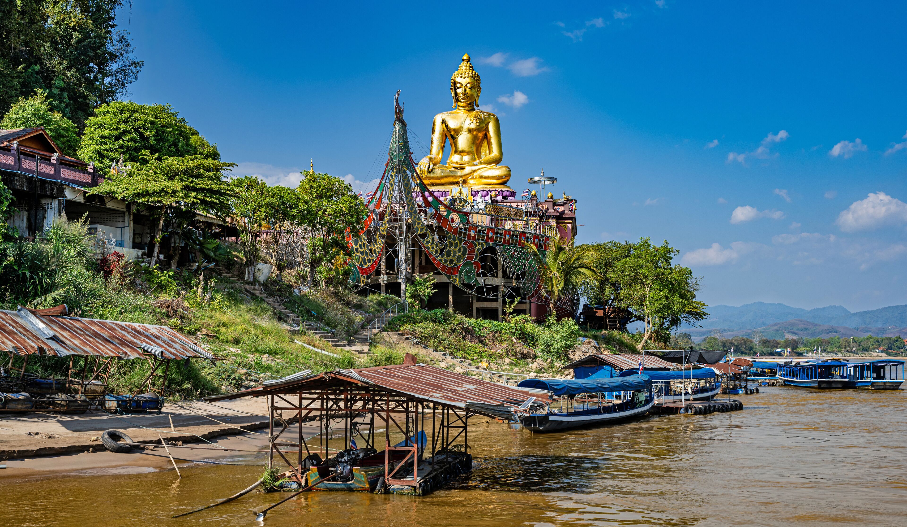 A large golden Buddha statue on a hill with boats docked in the water below.