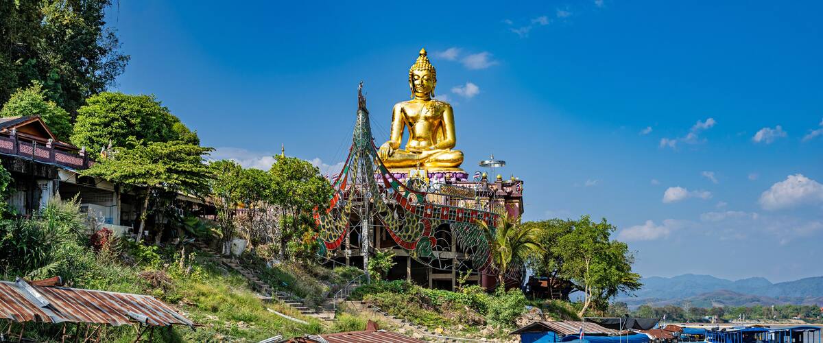 A large golden Buddha statue on a hill with boats docked in the water below.