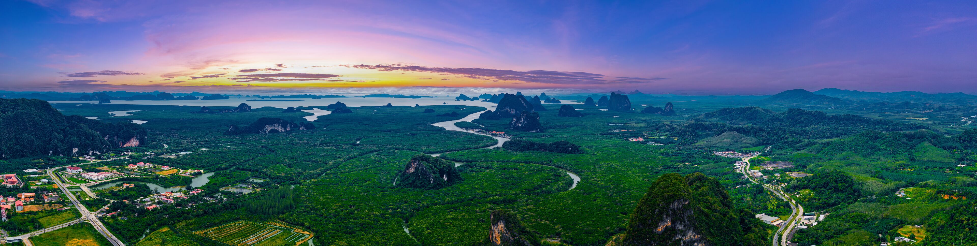 A lush green landscape with winding rivers, a variety of vegetation, and distant mountains under a colorful sunset sky.