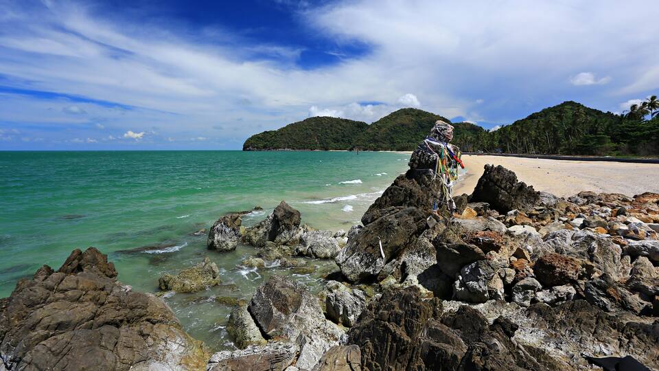 A rocky shoreline with clear turquoise water, a sandy beach, and a mountain in the background.
