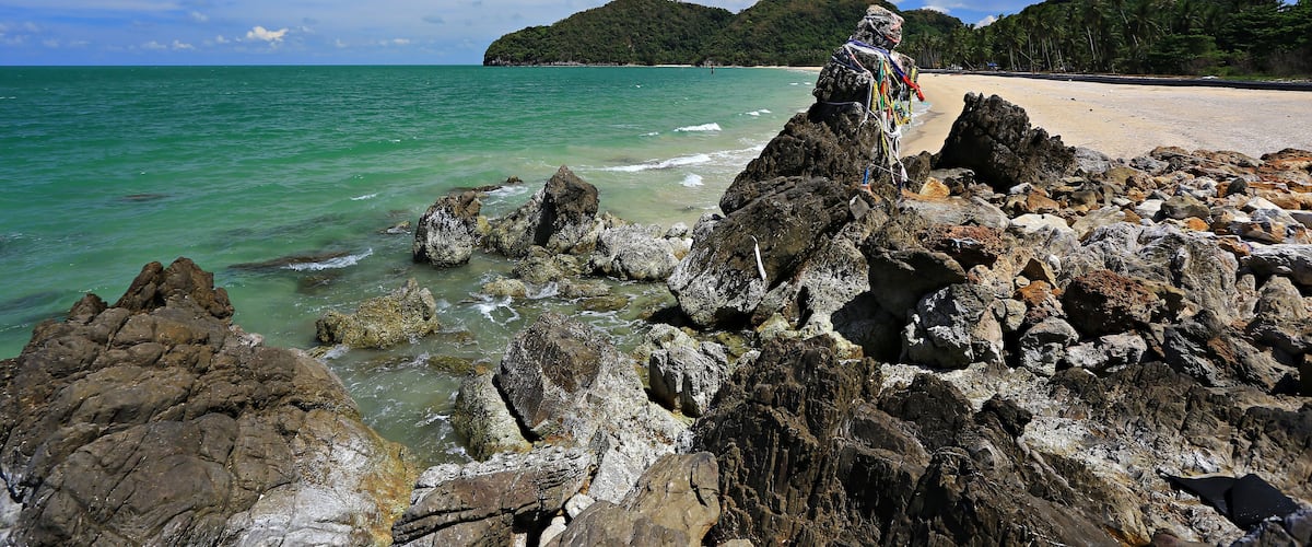 A rocky shoreline with clear turquoise water, a sandy beach, and a mountain in the background.
