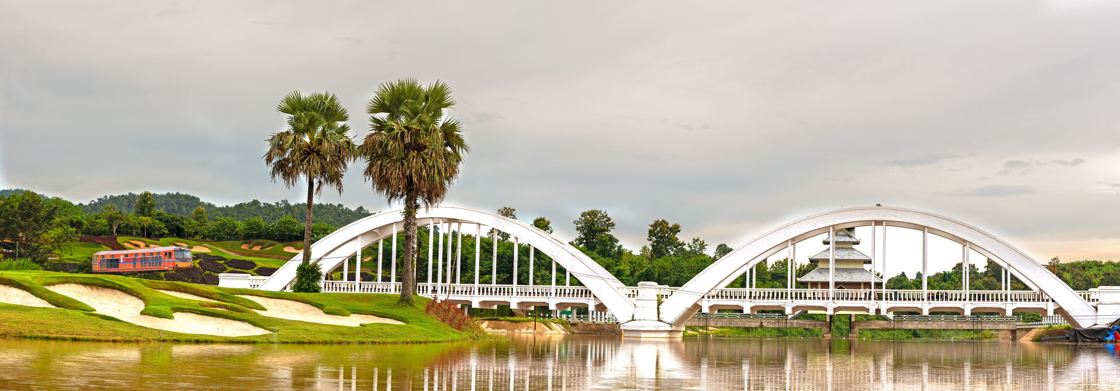 A white bridge with a pagoda-like structure on top, palm trees, and a red train on the tracks.
