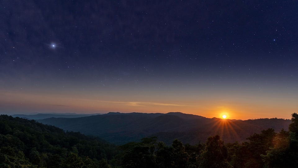 A starry night sky over a mountainous landscape with the sun setting on the horizon.