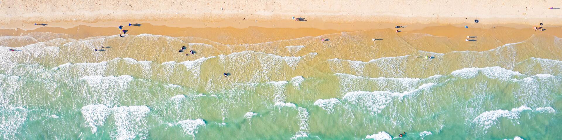 An aerial view of a beach with clear turquoise waters, a sandy shore, and dense green forest in the background.