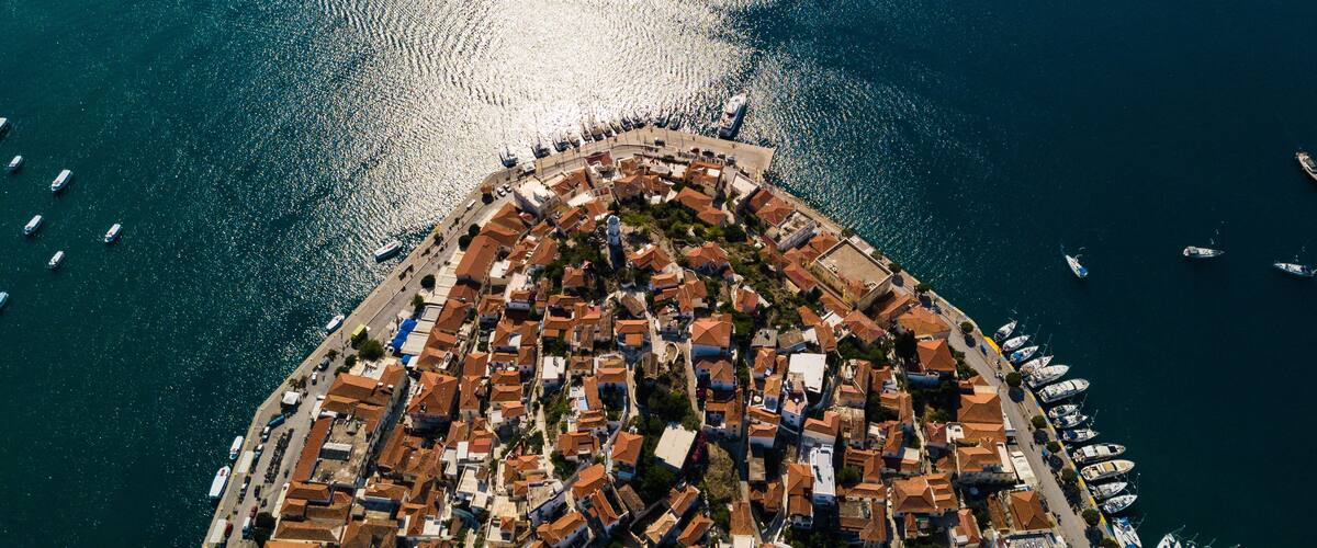 An aerial view of a coastal town with a dense cluster of buildings, a marina with numerous boats, and a clear view of the sea.