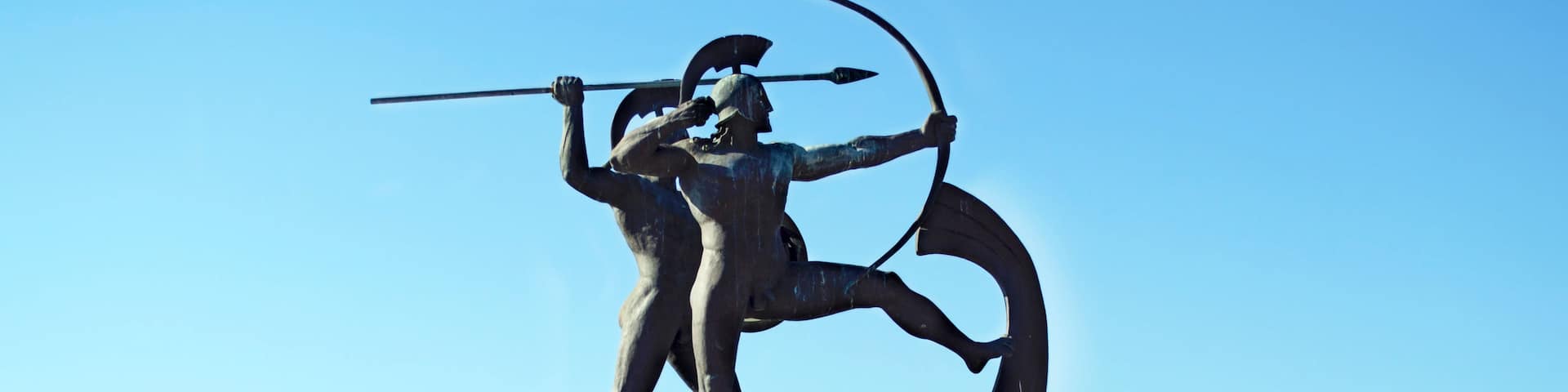 A bronze statue of a warrior with a spear and a curved sword, mounted on a pedestal with a clear sky and mountains in the background.