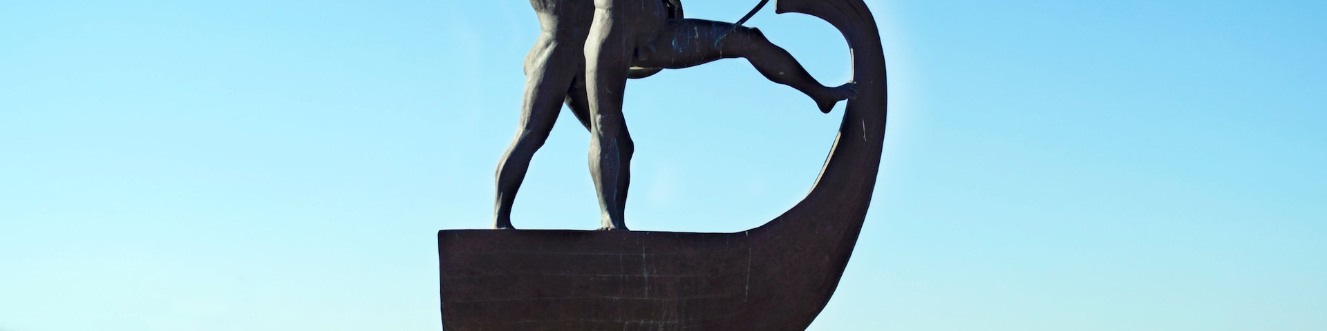Una estatua de bronce de un guerrero con una lanza y una espada curva, sobre un pedestal con cielo despejado y montañas al fondo.