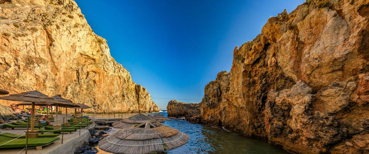 A coastal walkway with green cushioned benches and thatched umbrellas, leading to a rocky cliffside.