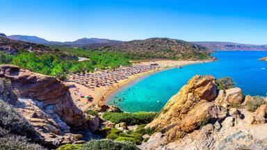 A coastal landscape with a beach, clear blue waters, rocky cliffs, and distant mountains.