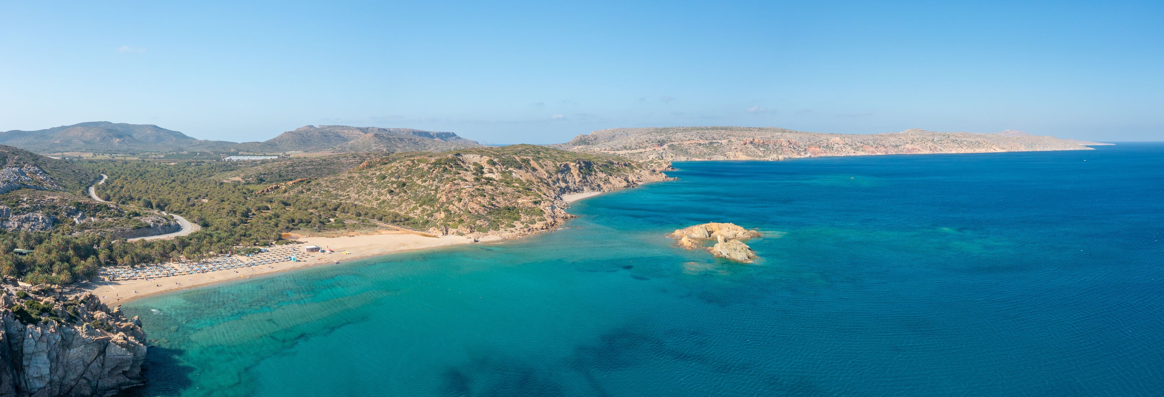 A coastal landscape with a beach, clear turquoise waters, rocky outcrops, and distant mountains.