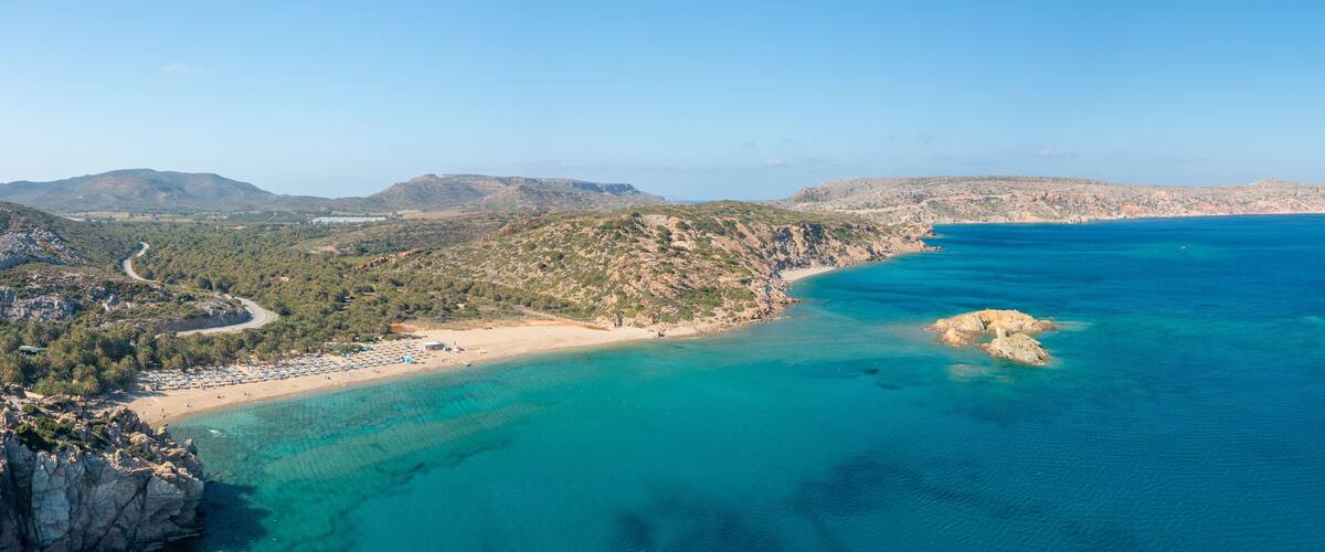 A coastal landscape with a beach, clear turquoise waters, rocky outcrops, and distant mountains.