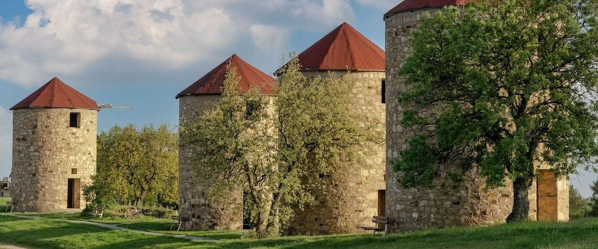 A row of stone towers with red roofs, surrounded by lush greenery and a clear blue sky.