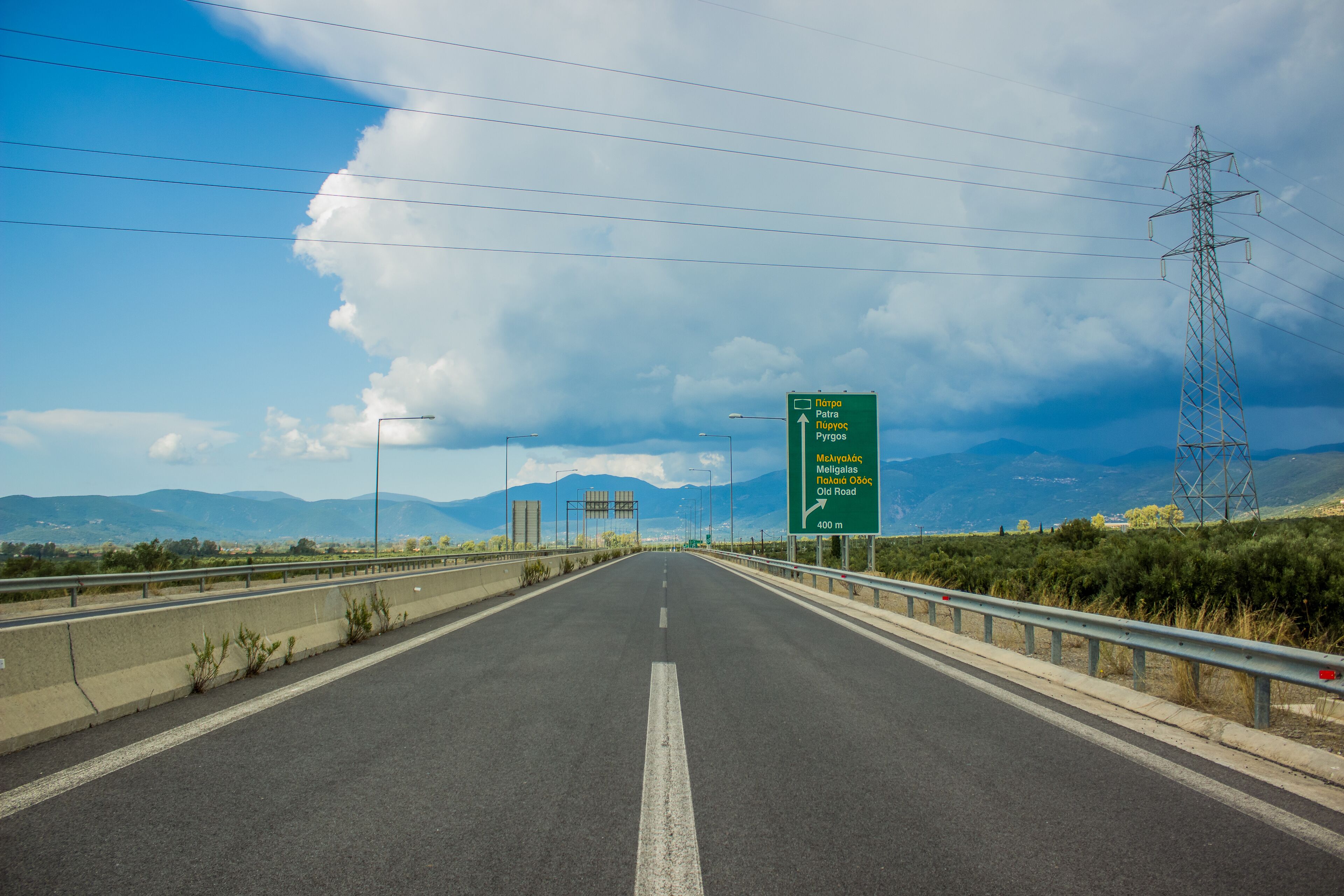 A highway with a green directional sign, guardrails, and power lines against a mountainous backdrop.