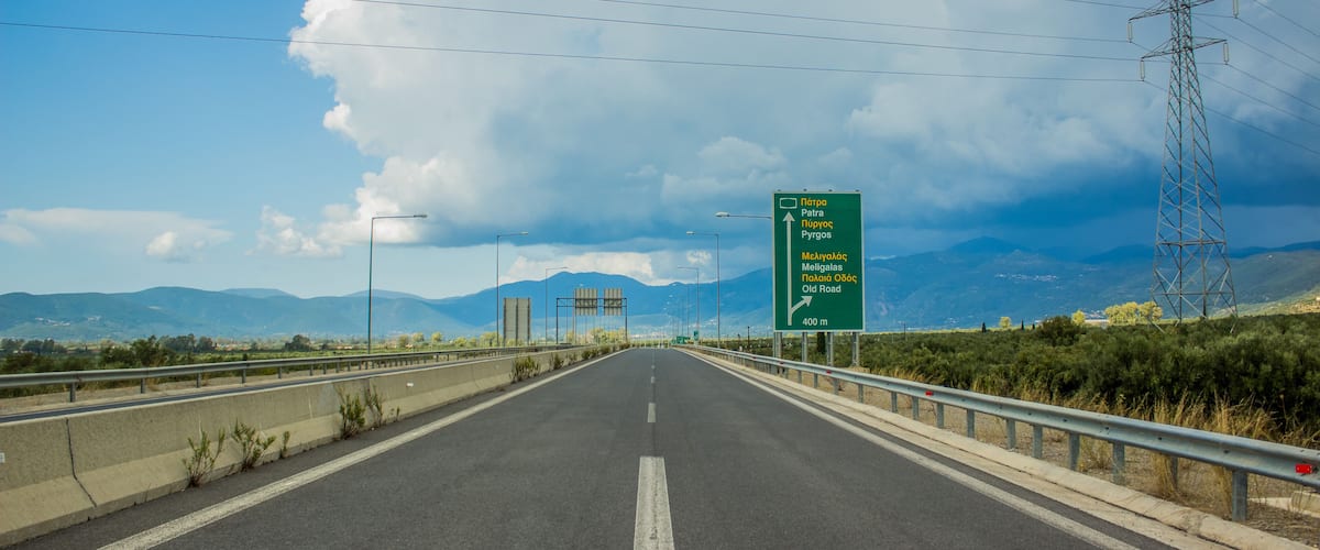 A highway with a green directional sign, guardrails, and power lines against a mountainous backdrop.