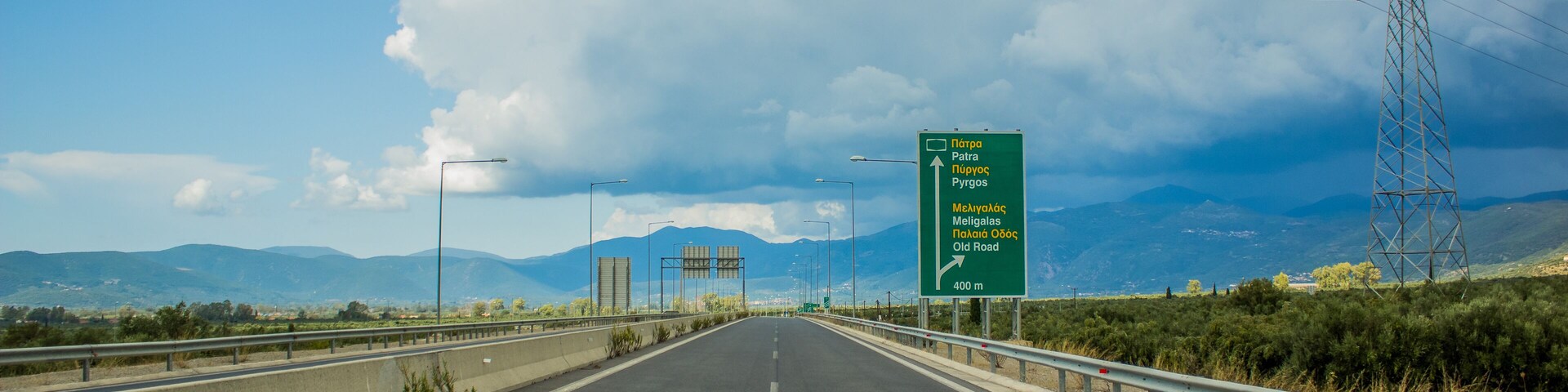 A highway with a green directional sign, guardrails, and power lines against a mountainous backdrop.