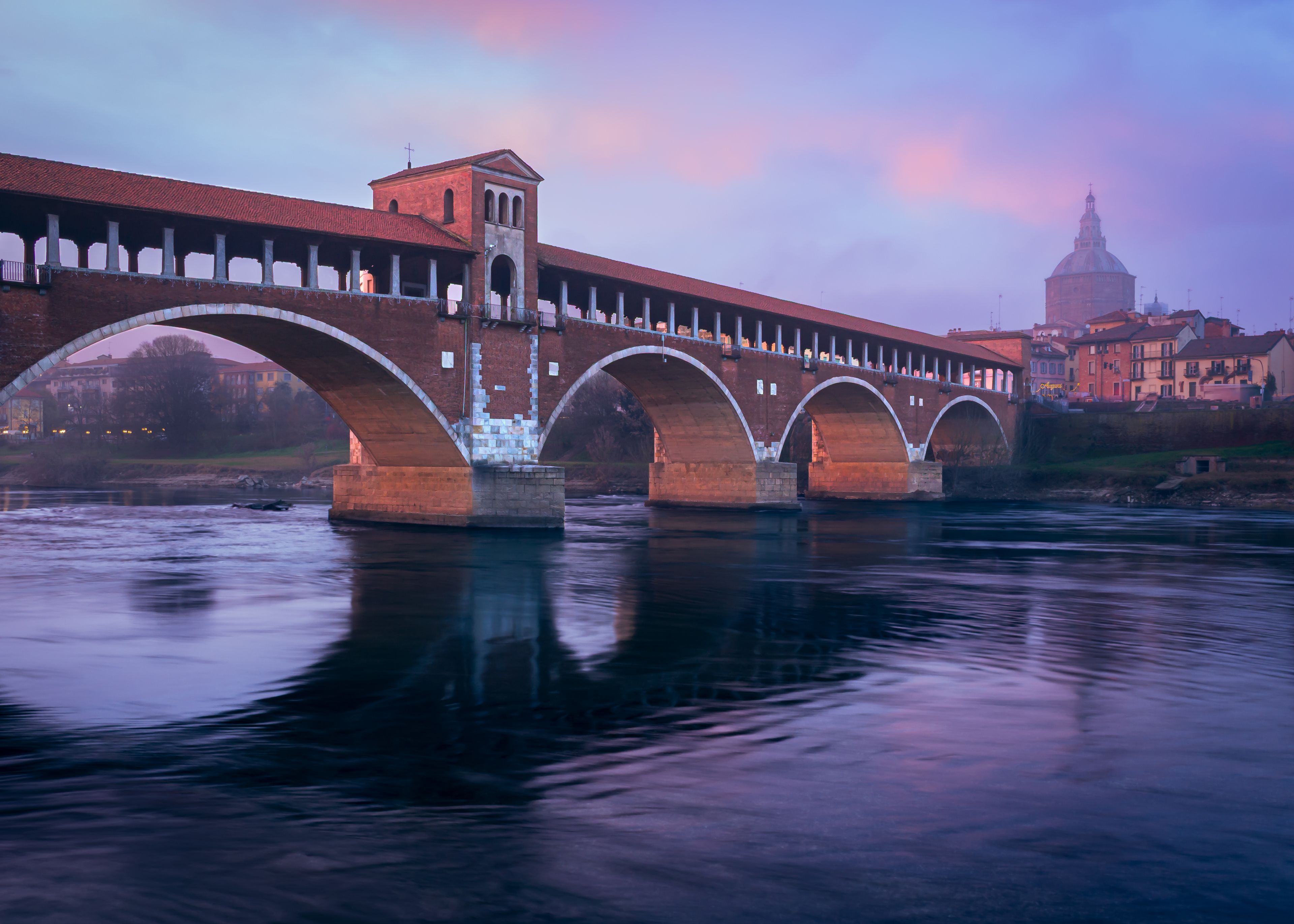 Un puente con varios arcos sobre un río, con edificios y una cúpula al fondo.