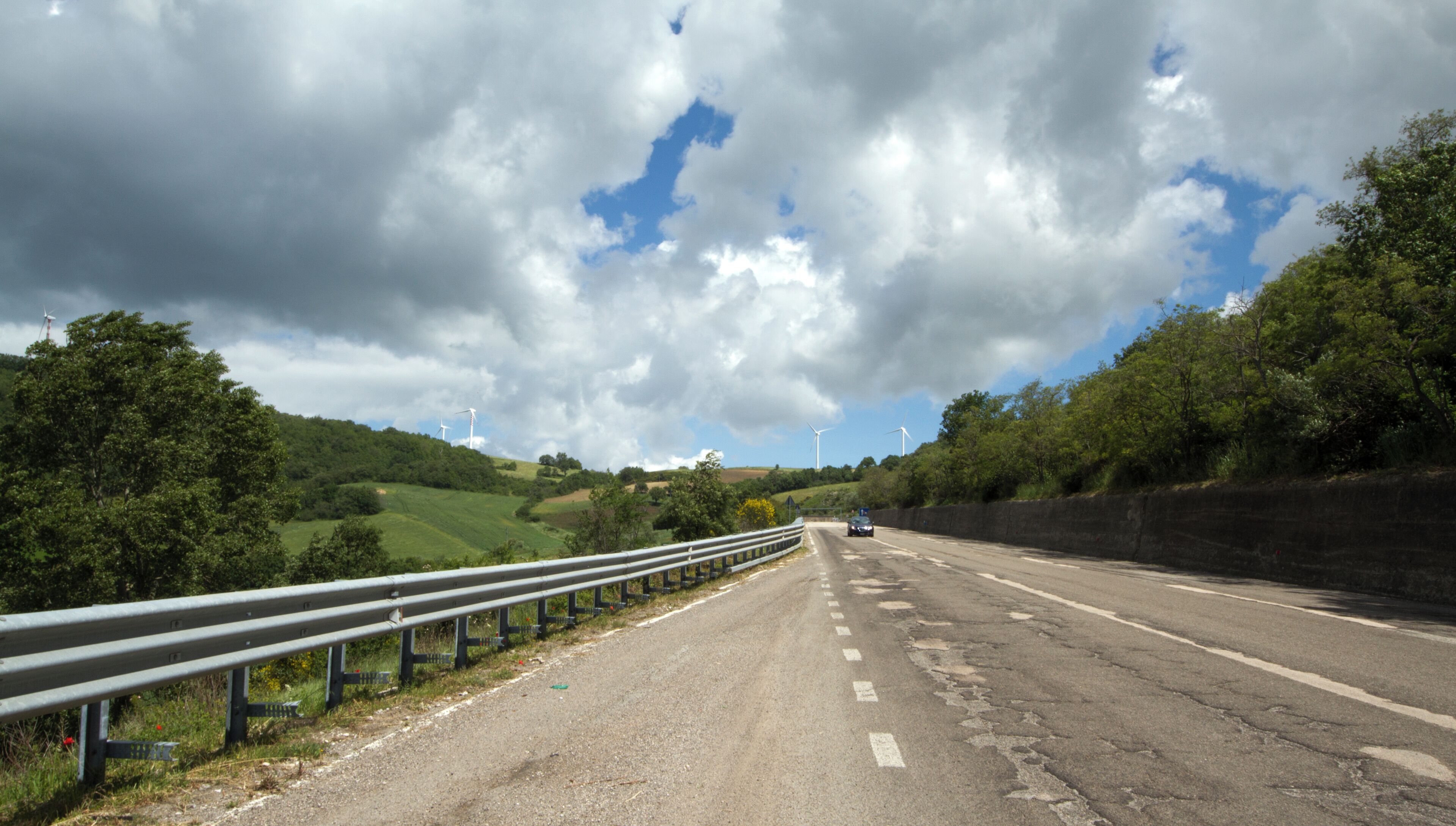 A rural road with wind turbines in the distance, surrounded by greenery and under a cloudy sky.