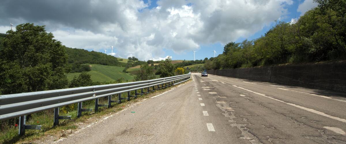 A rural road with wind turbines in the distance, surrounded by greenery and under a cloudy sky.