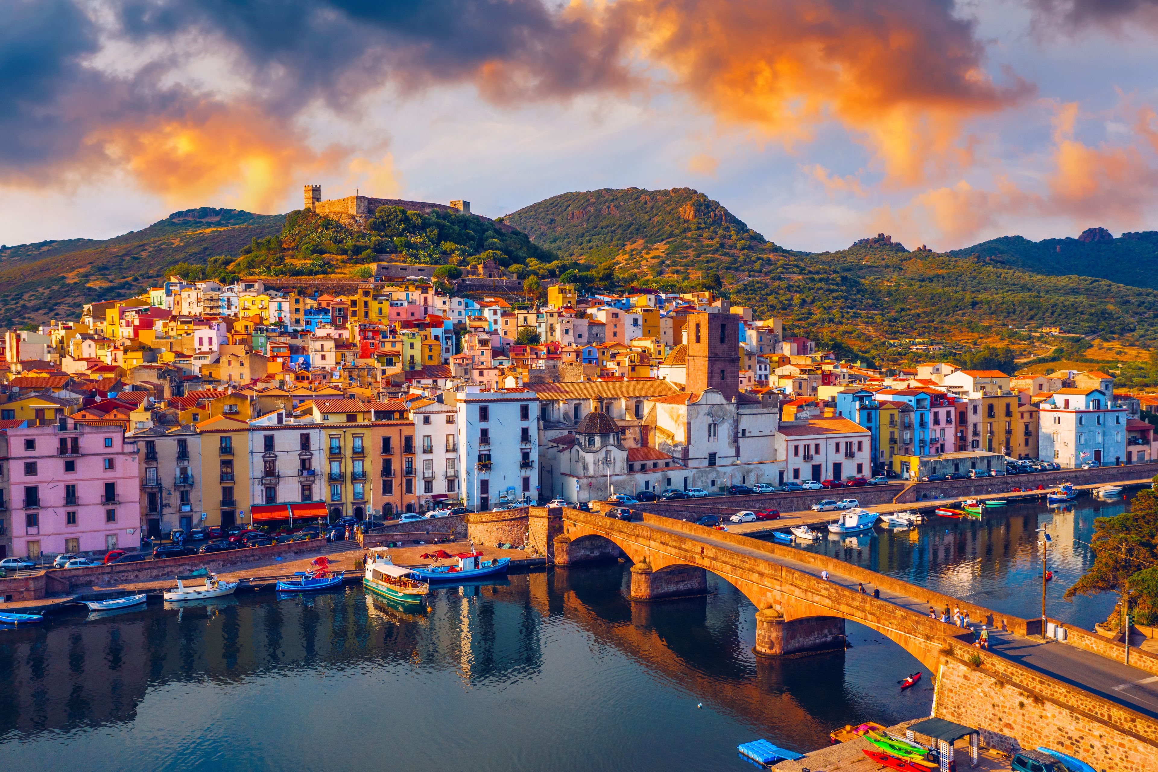 A coastal town with colorful buildings, a bridge over water, and hills in the background.