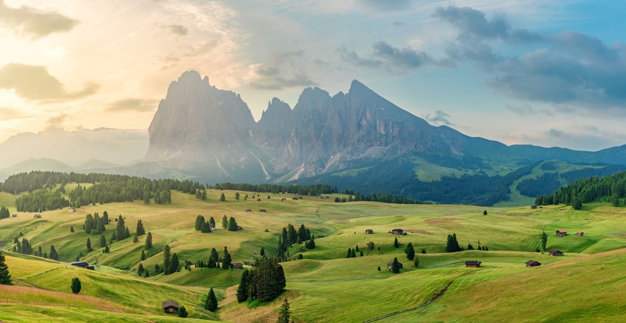 A mountainous landscape with green fields, scattered trees, and a prominent rocky peak.