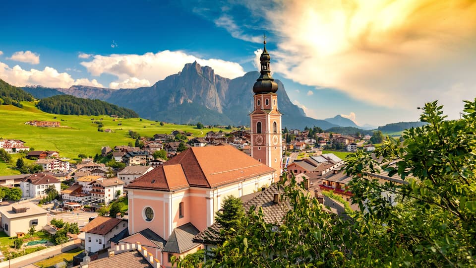 A town with a prominent church tower, lush green fields, and mountains in the background.