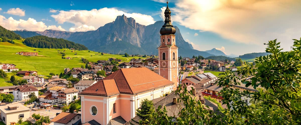 A town with a prominent church tower, lush green fields, and mountains in the background.