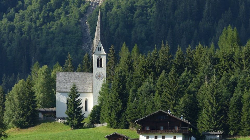 A white church with a clock tower surrounded by a forested mountain.