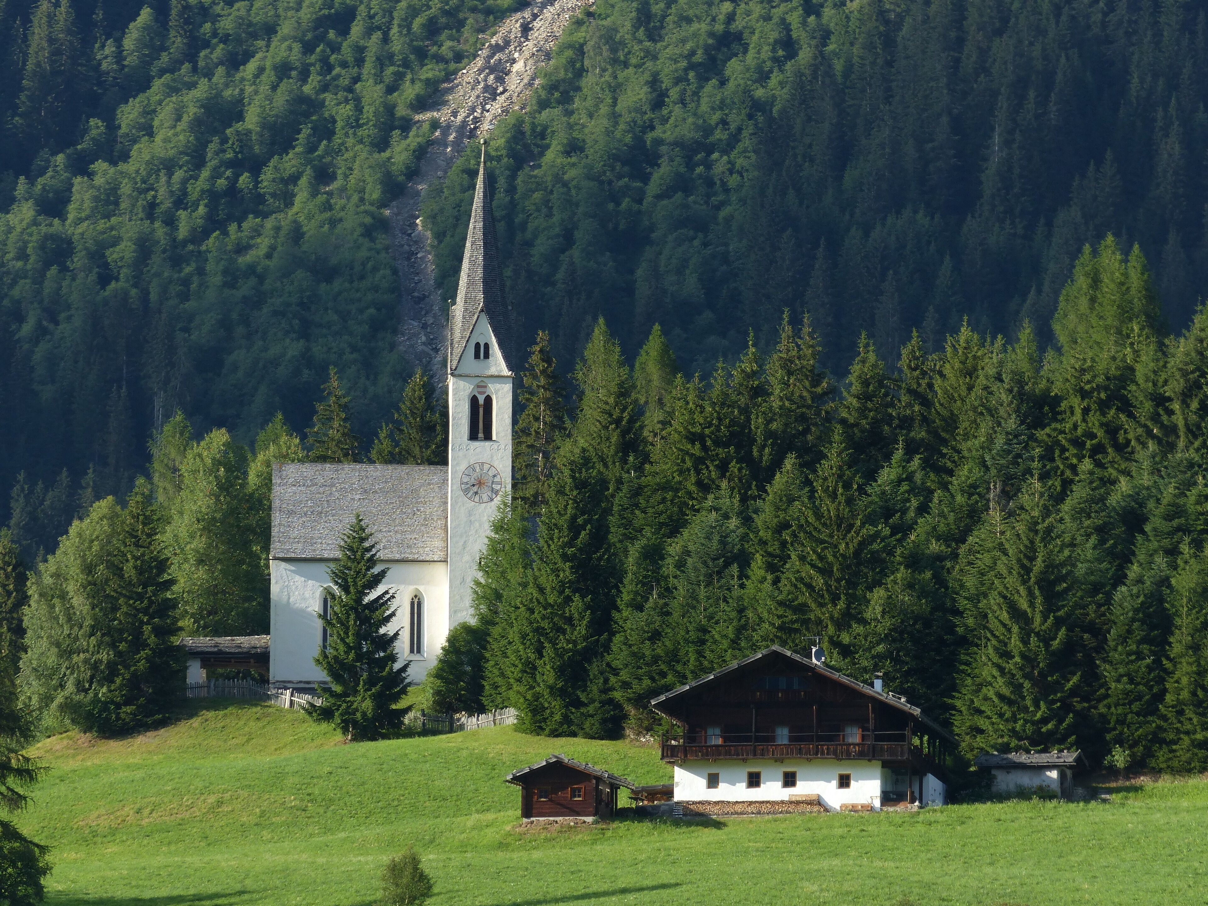 A white church with a clock tower surrounded by a forested mountain.