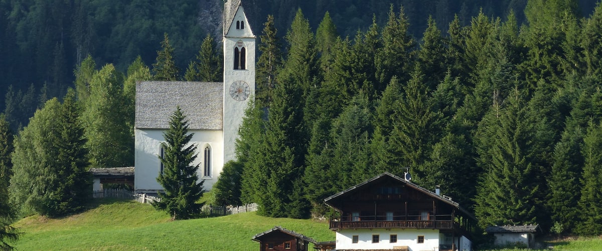 A white church with a clock tower surrounded by a forested mountain.