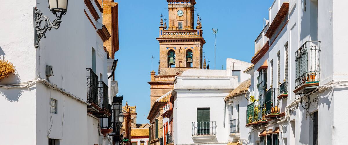 A narrow street with white buildings, a clock tower, and a clear blue sky.
