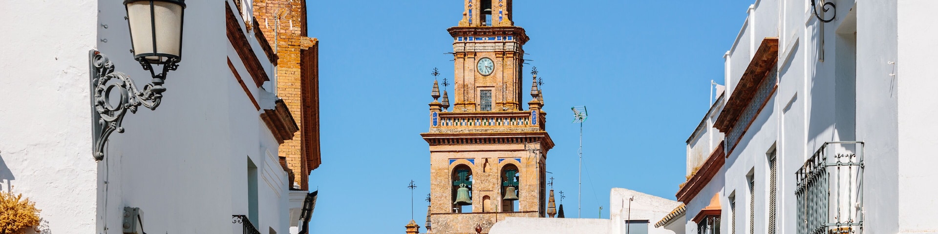 A narrow street with white buildings, a clock tower, and a clear blue sky.