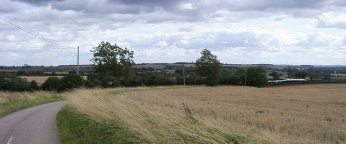 A rural road curving through a field with a cloudy sky.