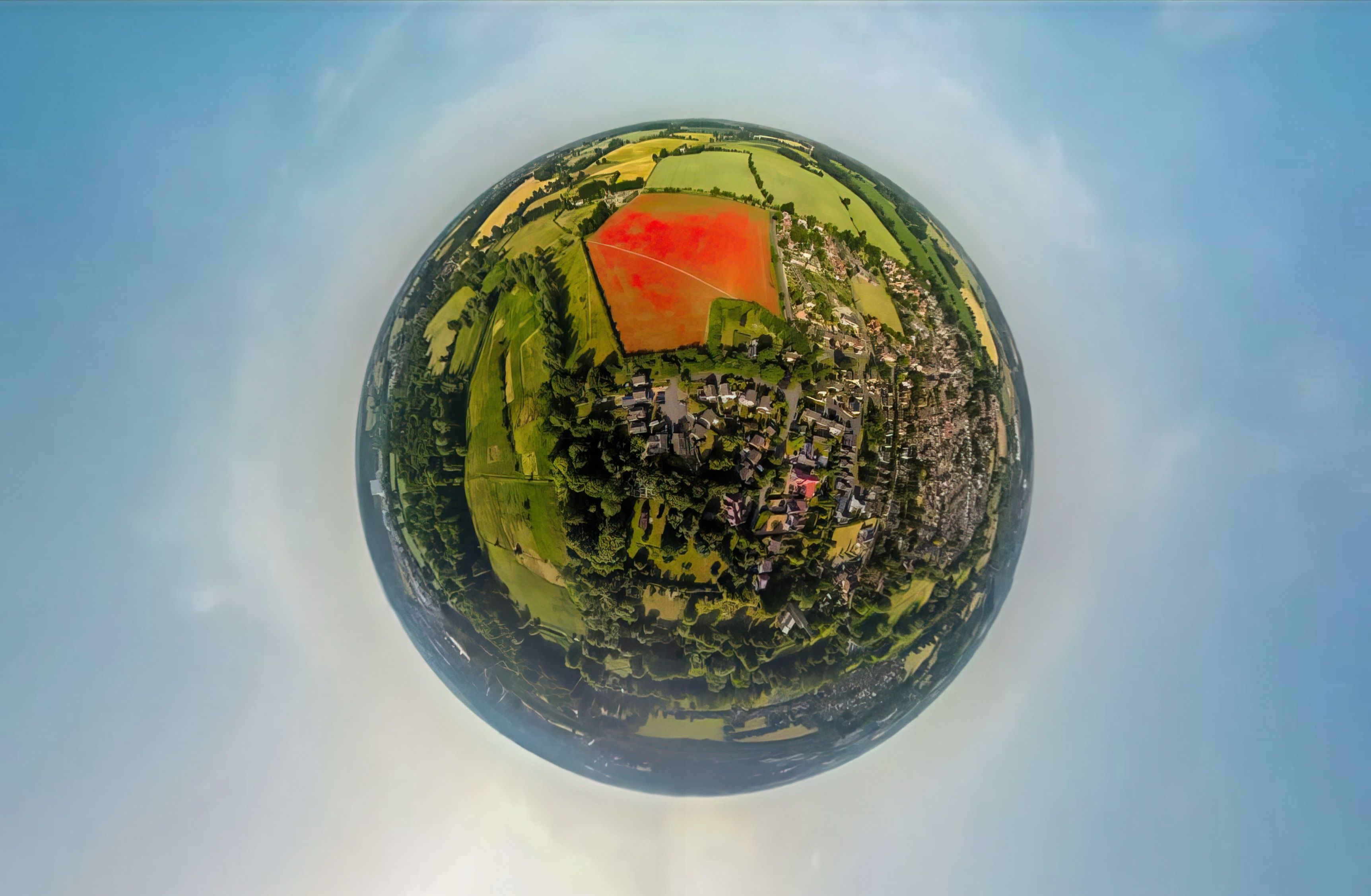 A spherical view of a landscape with a prominent red area, possibly a field or construction site, surrounded by green fields and scattered buildings.