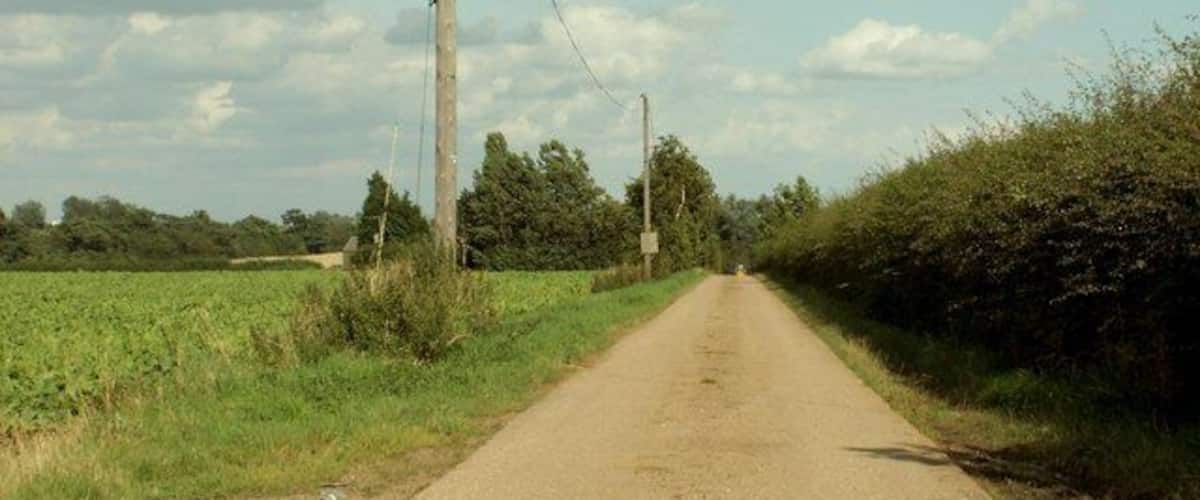 A rural road flanked by fields and hedgerows under a partly cloudy sky.