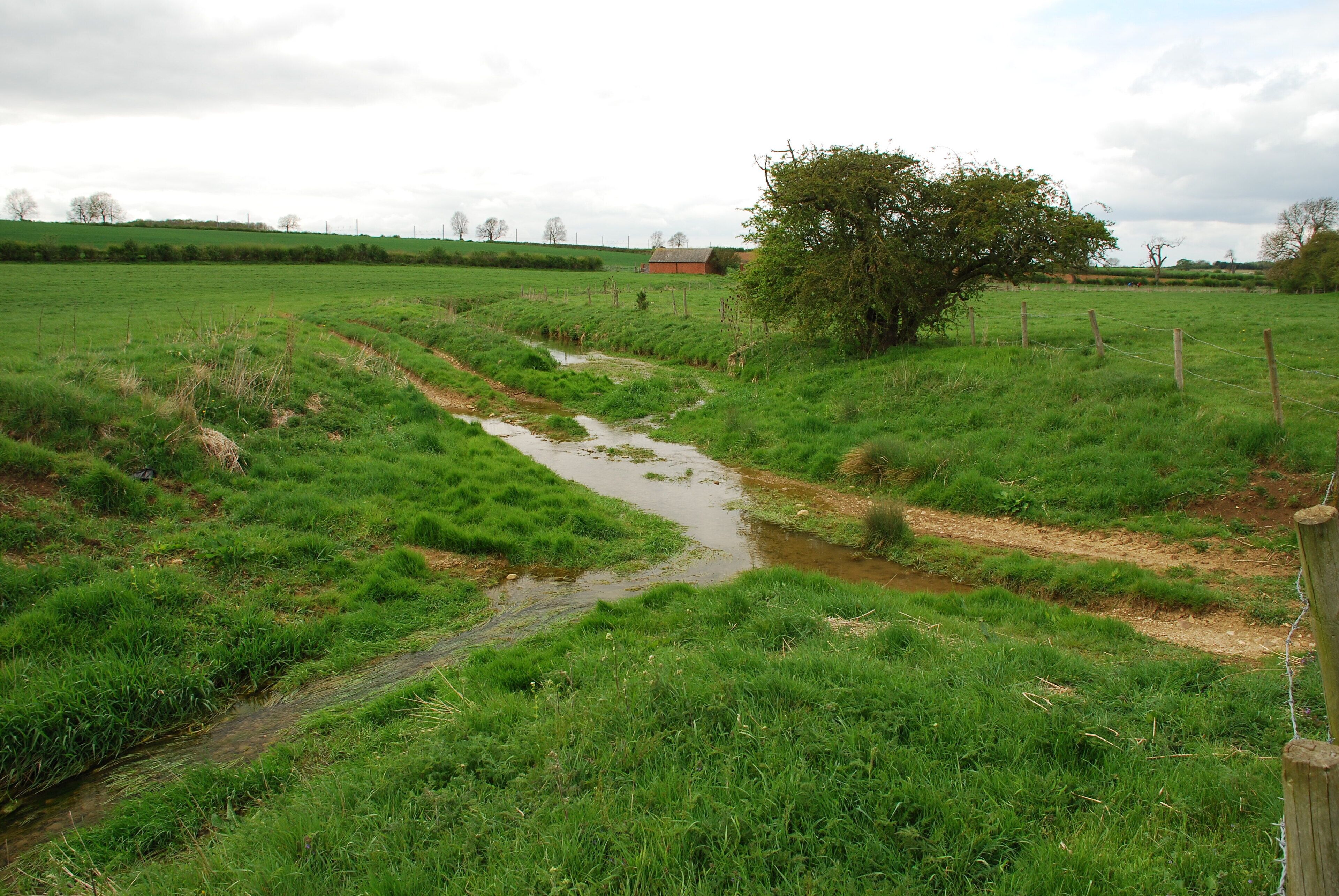 A rural landscape with a narrow stream, green fields, a wooden fence, and a distant red barn.