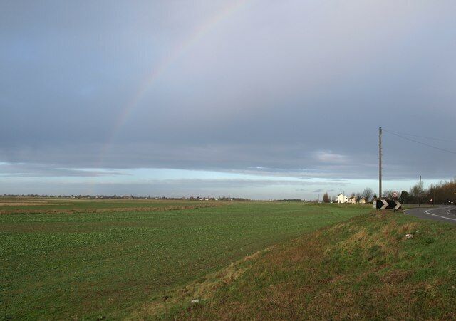 A rural landscape with a road curving to the right, a rainbow in the sky, and a field of crops.