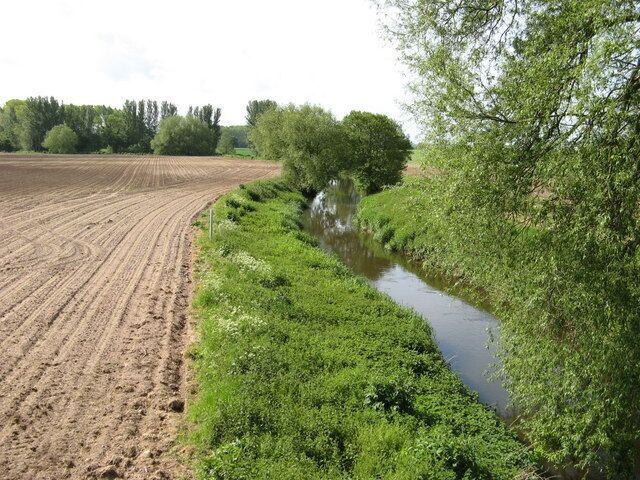 A river meandering through a field with a dirt path on one side and dense greenery on the other.