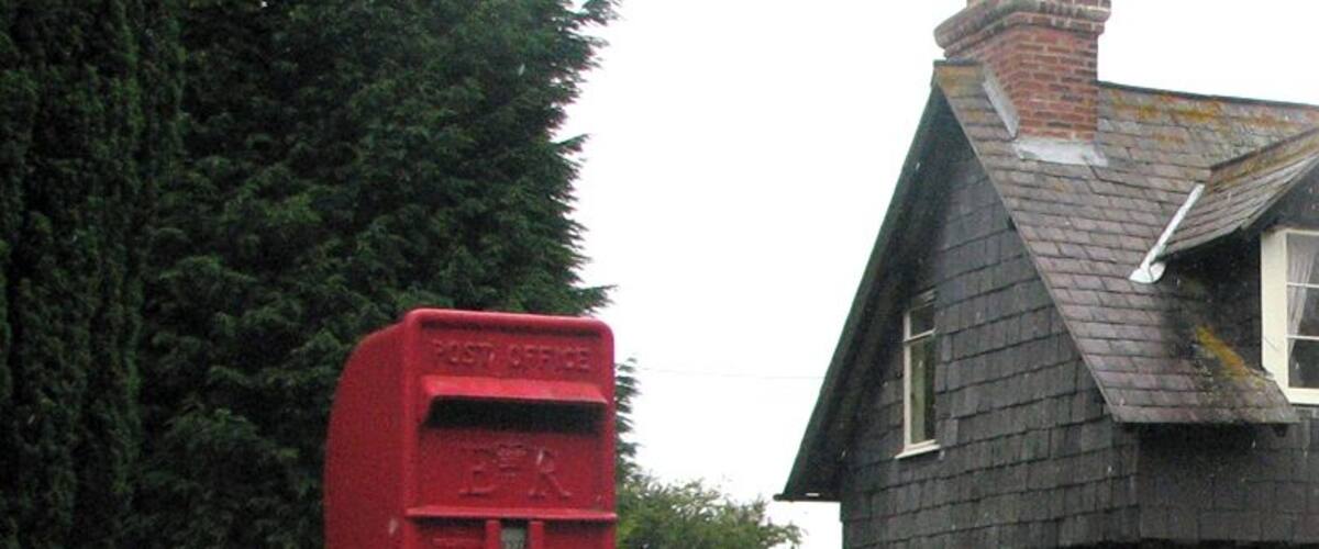 A red post office mailbox in a garden with purple flowers and a stone house in the background.