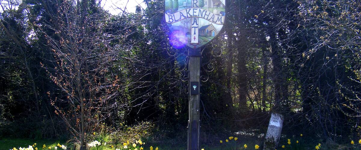 A signpost with 'Blakenham' written on it, surrounded by daffodils and trees.