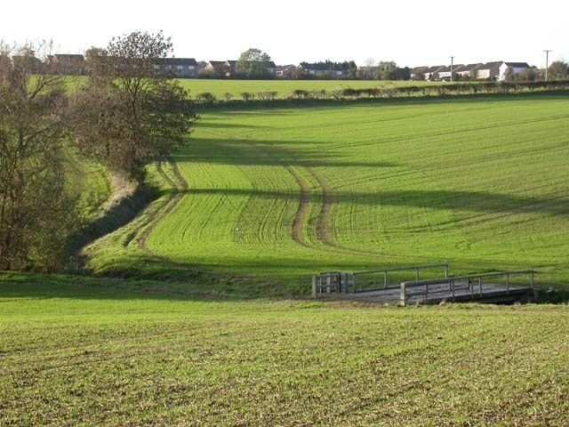 View towards Irchester A relatively new part of the village is visible. The footbridge at the bottom of the slope carries farm machinery, as well as walkers, across the brook.