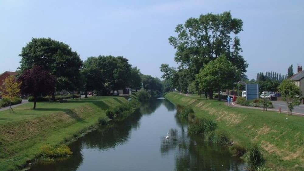 A calm river flanked by trees and grassy banks, with a clear sky above.