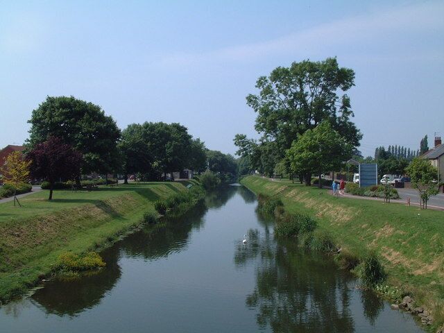 A calm river flanked by trees and grassy banks, with a clear sky above.