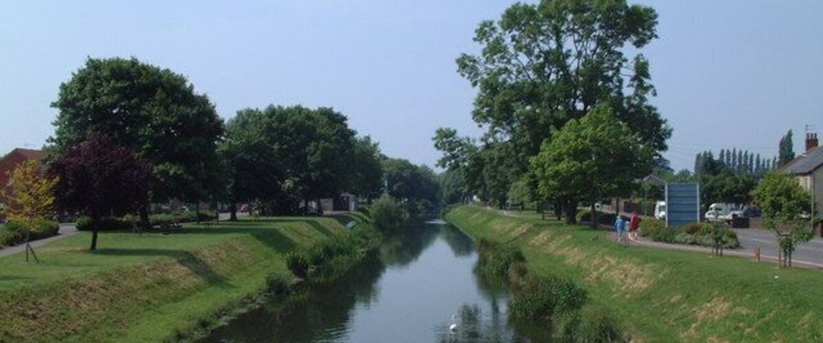 A calm river flanked by trees and grassy banks, with a clear sky above.