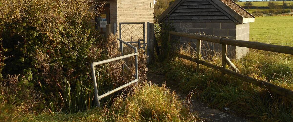 A rural scene with two small stone buildings, a wooden gate, and a grassy field.