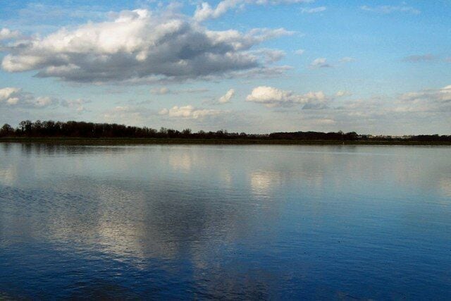 Um lago calmo refletindo o céu e árvores ao longe.