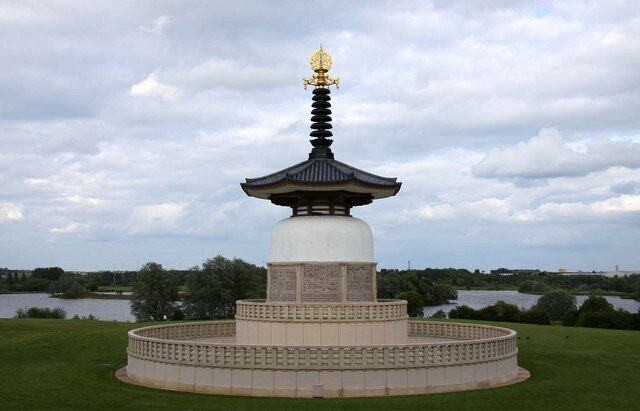 Monument d'estil pagoda amb part superior daurada, envoltat per una base circular de pedra, davant d'un cel nuvolós i un llac al fons.