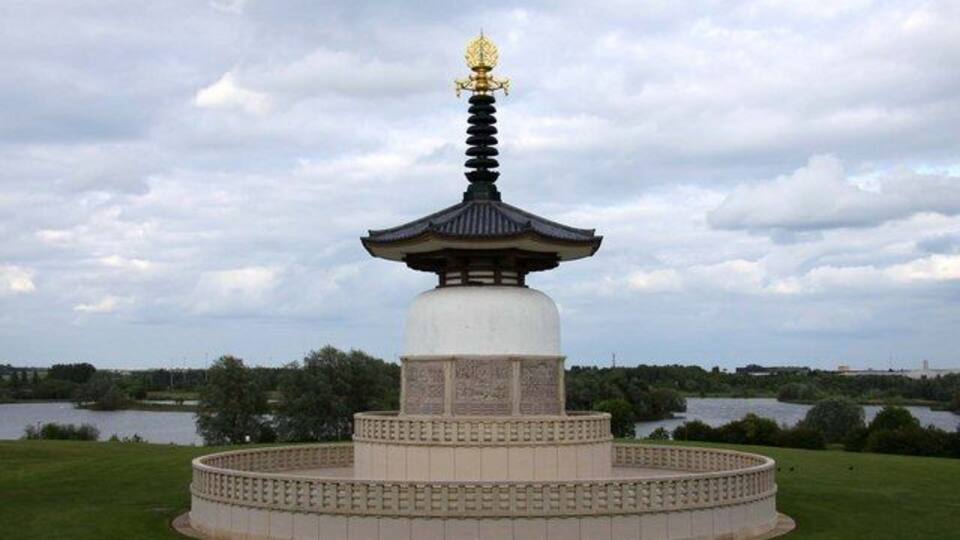 Monument d'estil pagoda amb part superior daurada, envoltat per una base circular de pedra, davant d'un cel nuvolós i un llac al fons.
