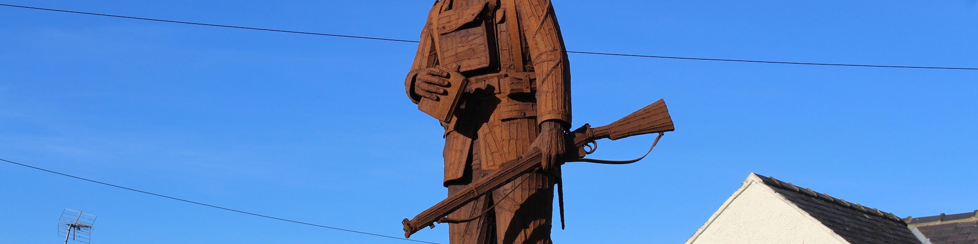 A bronze statue of a soldier holding a rifle, standing on a pedestal with a wreath, in front of a white house with a sloped roof.