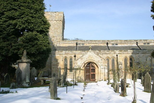 A historic church with a snow-covered graveyard and a large tree to the left.