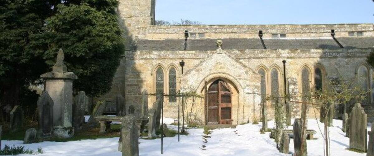 A historic church with a snow-covered graveyard and a large tree to the left.